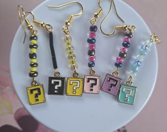 Può includere: Una collezione di orecchini fatti a mano con ganci color oro. Ogni orecchino presenta un ciondolo quadrato con un punto interrogativo e accenti di perline colorate. I ciondoli sono gialli, neri, rosa, viola e turchese.