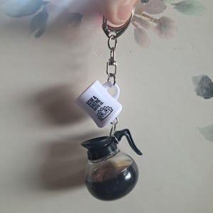 May include: A coffee-themed keychain featuring a miniature white mug and a glass coffee pot. The mug has the text "Rise & Shine Coffee Co." and a sun graphic. The coffee pot is filled with dark liquid.