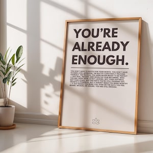 May include: A framed print with the text "YOU'RE ALREADY ENOUGH" in a bold, black font. Below the title is a longer paragraph in a smaller font. The frame is a light brown colour, and the background is a neutral beige.