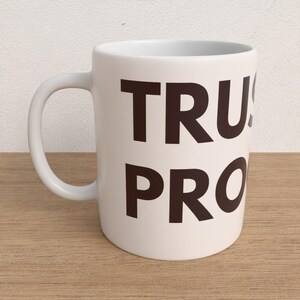 Op de afbeelding: Witte keramische mok met een gebogen handvat. De mok heeft de woorden "TRUST PRO" in grote, donkerbruine blokletters. De mok staat op een lichtbruin houten oppervlak tegen een witte achtergrond.