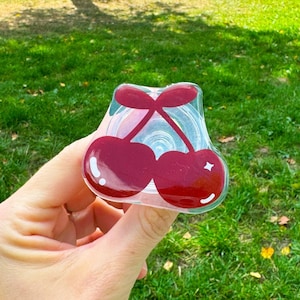 May include: A clear plastic cherry-shaped dispenser with red cherries inside. The dispenser is being held in a hand.