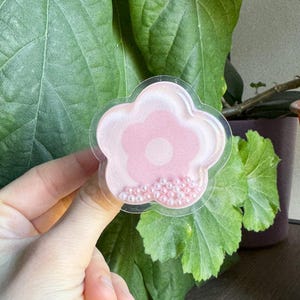 Può includere: Un grip per telefono rosa a forma di fiore con una custodia in plastica trasparente e uno strato di perline bianche perlate attorno al bordo.