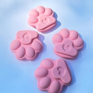 Peut inclure: Quatre moules en silicone roses en forme de pattes, chacun avec un détail en forme de cœur au centre. Les moules sont disposés sur une surface bleu clair, avec des ombres douces. Le design est simple et mignon.