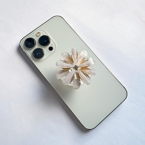 Könnte beinhalten: Ein hellgrünes Smartphone mit einem floralen Handyhalter. Der Handyhalter hat eine transparente Basis mit rosa- und goldfarbenen Blütenblättern, die kreisförmig angeordnet sind, und einen kleinen silbernen Edelstein in der Mitte.