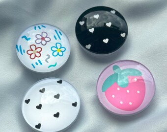 Può includere: Quattro oggetti decorativi rotondi con diversi disegni. Uno ha fiori, un altro cuori bianchi su nero, un terzo cuori neri su bianco e l'ultimo una fragola rosa con un gambo verde.