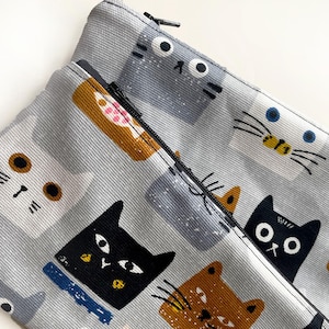 Op de afbeelding: Een grijze ritszak met een herhalend patroon van katten in verschillende kleuren en stijlen. De katten zijn getekend in een cartoon-stijl met eenvoudige kenmerken. De zak is gemaakt van een zachte, textuurrijke stof.