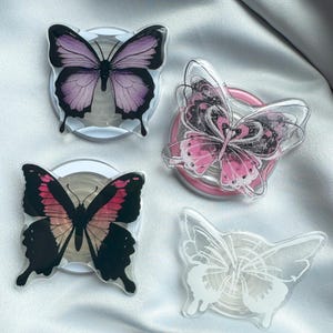 May include: Four butterfly-shaped phone grips in various colors. One has purple wings, another pink, and a third has black and pink wings. The fourth is white. Each grip is attached to a round base.