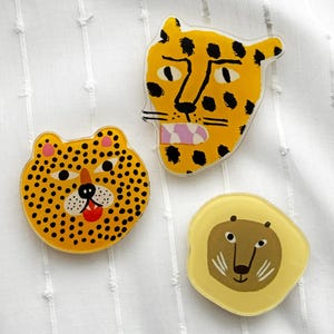 May include: Three animal-themed badges: a leopard, a cheetah, and a lion. The leopard badge is yellow with black spots and a pink mouth. The cheetah badge is yellow with black spots and a red tongue. The lion badge is yellow with a brown face.
