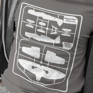 May include: Gray t-shirt featuring a white graphic of a model airplane kit. The kit design includes the text "Superior Model Co." and "Spitfire Mk. IXe Scale 1:48". The shirt is a casual wear item.
