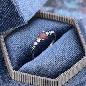 May include: A silver ring with a red square gemstone surrounded by smaller clear gemstones.