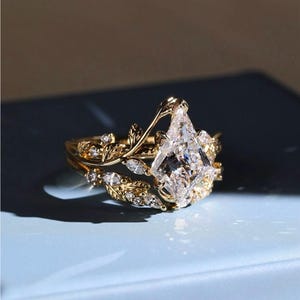 Peut inclure: Un ensemble de bagues dorées avec une grosse pierre précieuse claire en forme de cerf-volant. La bague est ornée de feuilles et de diamants, créant un design élégant et inspiré de la nature. L'ensemble comprend une alliance assortie.
