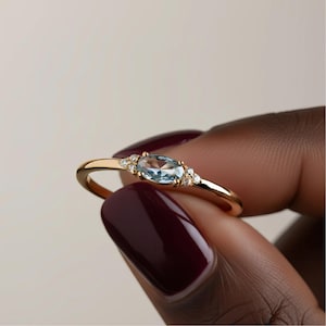 Könnte beinhalten: Ein goldener Ring mit einem hellblauen, ovalen Edelstein und kleineren, klaren Steinen. Der Ring hat ein zierliches Band. Der Ring wird vor einem neutralen Hintergrund gehalten.