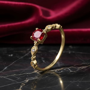 Puede incluir: Un anillo de oro con una gran gema roja y redonda como pieza central. La banda presenta un diseño delicado con pequeñas piedras claras. El anillo está sobre un fondo oscuro con una tela roja.