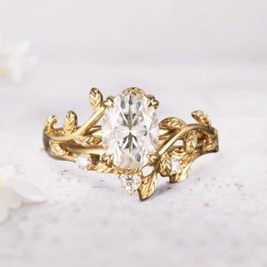 May include: A gold engagement ring with an oval-cut diamond center stone. The ring features a leaf and vine design with small accent diamonds. The band and setting are gold-colored, creating a nature-inspired aesthetic.