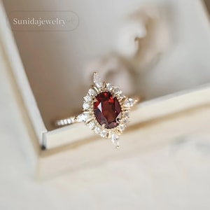 May include: A gold ring with a large, oval, deep red gemstone, encircled by smaller white gemstones. The ring has a fine band and a halo design. It is presented in a white box. The text "Sunidajewelry" is in the upper left corner.