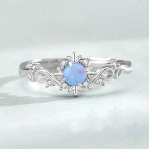 May include: A silver ring featuring a snowflake design with a light blue opal center stone. The ring is adorned with small, clear gemstones and has a delicate, swirling band. The ring is a piece of jewelry.