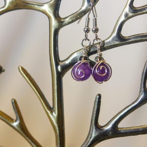 May include: A pair of purple amethyst earrings with silver wire wrapping, hanging from a decorative bronze tree with small bird figurines. The earrings have a round shape and a spiral design.