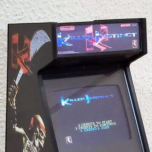 Op de afbeelding: Een zwarte Killer Instinct arcadekast met een scherm waarop de titel van het spel wordt weergegeven. De kast heeft een skeletafbeelding, blauwe joysticks en de logo's van Nintendo en Midway. De tekst "2 Credits to Continue" is zichtbaar op het scherm.