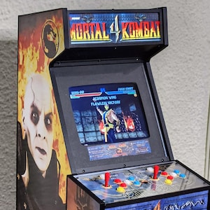 Puede incluir: Una máquina recreativa negra de Mortal Kombat 4 con una pantalla que muestra el juego. El mueble presenta ilustraciones de un personaje y el logotipo del juego. Tiene botones rojos, azules y blancos y un joystick para jugar.