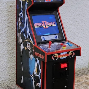 Op de afbeelding: Miniatuur arcadekast met het spel Mortal Kombat II. De kast is zwart met rode randen, een joystick, knoppen en een scherm met de titel van het spel. De zijkanten tonen karakterafbeeldingen.