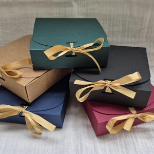 Pode incluir: Uma cole&ccedil;&atilde;o de caixas de presente coloridas em v&aacute;rias tonalidades, incluindo verde, castanho, azul, preto e bord&ocirc;. Cada caixa &eacute; amarrada com uma fita dourada. As caixas s&atilde;o quadradas com tampa curva.