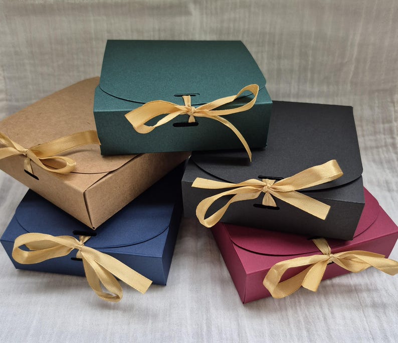 Pode incluir: Uma cole&ccedil;&atilde;o de caixas de presente coloridas em v&aacute;rias tonalidades, incluindo verde, castanho, azul, preto e bord&ocirc;. Cada caixa &eacute; amarrada com uma fita dourada. As caixas s&atilde;o quadradas com tampa curva.