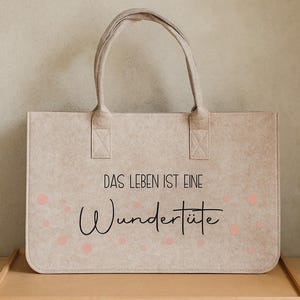 Könnte beinhalten: Beige Tragetasche mit rechteckiger Form und zwei Griffen. Die Tasche trägt den Text "DAS LEBEN IST EINE Wundertüte" in Schwarz mit rosa Punkten. Die Tasche ist aus einem filzartigen Material gefertigt.