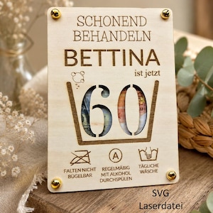 以下が含まれることがあります： 「SCHONEND BEHANDELN BETTINA ist jetzt 60」の文字が入った木製プレート。数字の60が切り抜かれており、背後の内容が見える。プレートの四隅には金色のネジがあり、ケアマークと「SVG Laserdatei」の文字も含まれています。