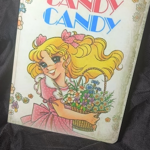 Puede incluir: Portada de libro vintage "Candy Candy" con una niña rubia con lazos rosas y un vestido rosa, sosteniendo una cesta de flores. El título está en rojo y azul. La portada tiene un borde floral y una ilustración de dibujos animados.