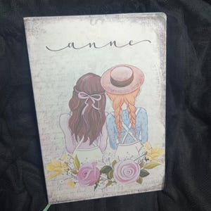 Könnte beinhalten: Ein Notizbuch im Vintage-Stil mit floralem Design und zwei Mädchen mit braunen und blonden Haaren. Das Notizbuch trägt den Namen "anne" auf der Vorderseite.