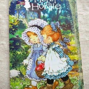Könnte beinhalten: Ein rechteckiger Buchumschlag mit dem Text Holly Hobbie oben. Die Illustration zeigt zwei Kinder in Kleidung im Vintage-Stil, eines mit einer blau gepunkteten Haube und das andere mit einer blauen Schleife, die einen Korb mit roten Äpfeln tragen.