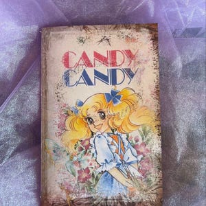 May include: A vintage-style book cover featuring a cartoon girl with blonde hair and a blue dress. The title "CANDY CANDY" is printed in bold letters on the cover.