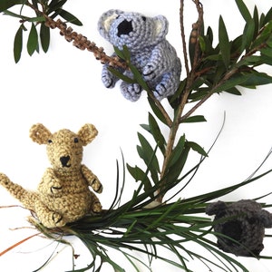 Op de afbeelding: Drie gehaakte knuffels: een grijze koala, een bruine kangoeroe en een zwart-grijze wombat. De dieren zitten op een bed van groen gras.