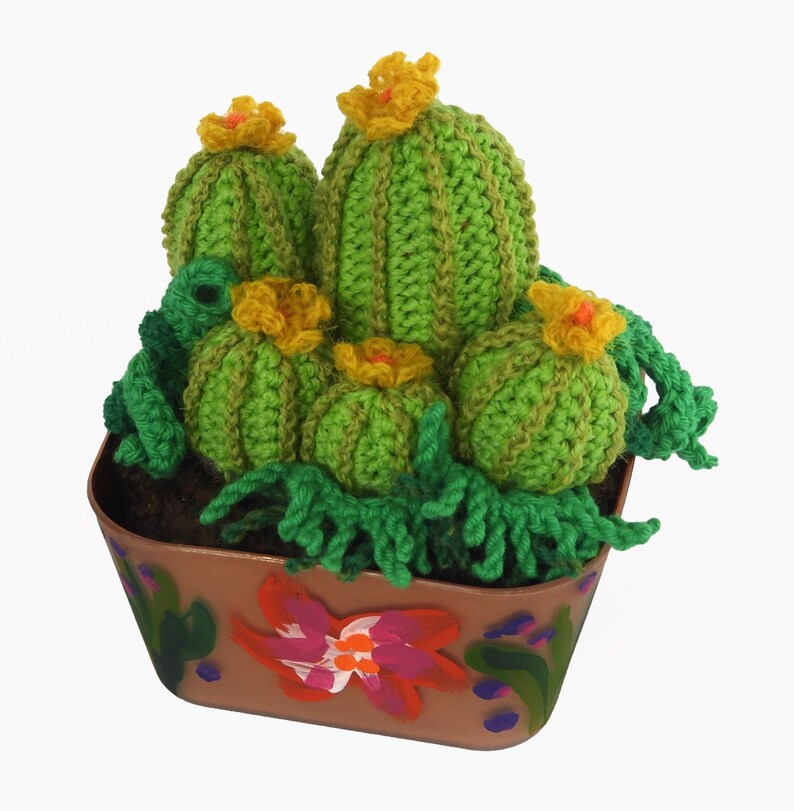 May include: A handcrafted crochet cactus arrangement in a brown painted pot. The cacti are various sizes, in shades of green, with yellow flowers. The pot is decorated with painted flowers and foliage.