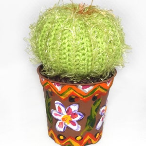 May include: A handmade, decorative cactus in a painted pot. The cactus is a round, light green yarn ball with fuzzy strands. The brown pot is adorned with hand-painted flowers and geometric patterns in orange, yellow, and white.