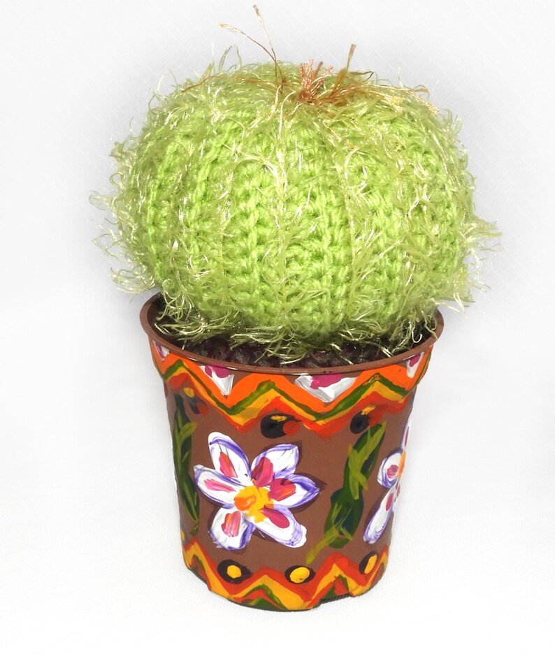 May include: A handmade, decorative cactus in a painted pot. The cactus is a round, light green yarn ball with fuzzy strands. The brown pot is adorned with hand-painted flowers and geometric patterns in orange, yellow, and white.