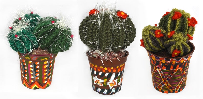 May include: Three handmade crochet cactus plants in decorative pots. The cacti are various shades of green with white accents and red flower details. The pots are brown with colorful, hand-painted designs, including geometric patterns and floral motifs.