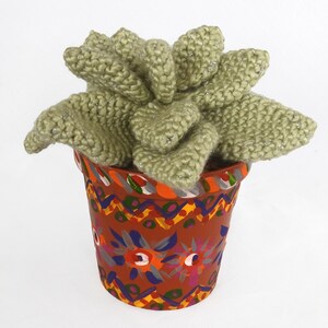May include: A handmade, knitted succulent plant in a decorative terracotta pot. The plant is a soft green color with silver thread accents. The pot is painted with colorful floral and geometric patterns.