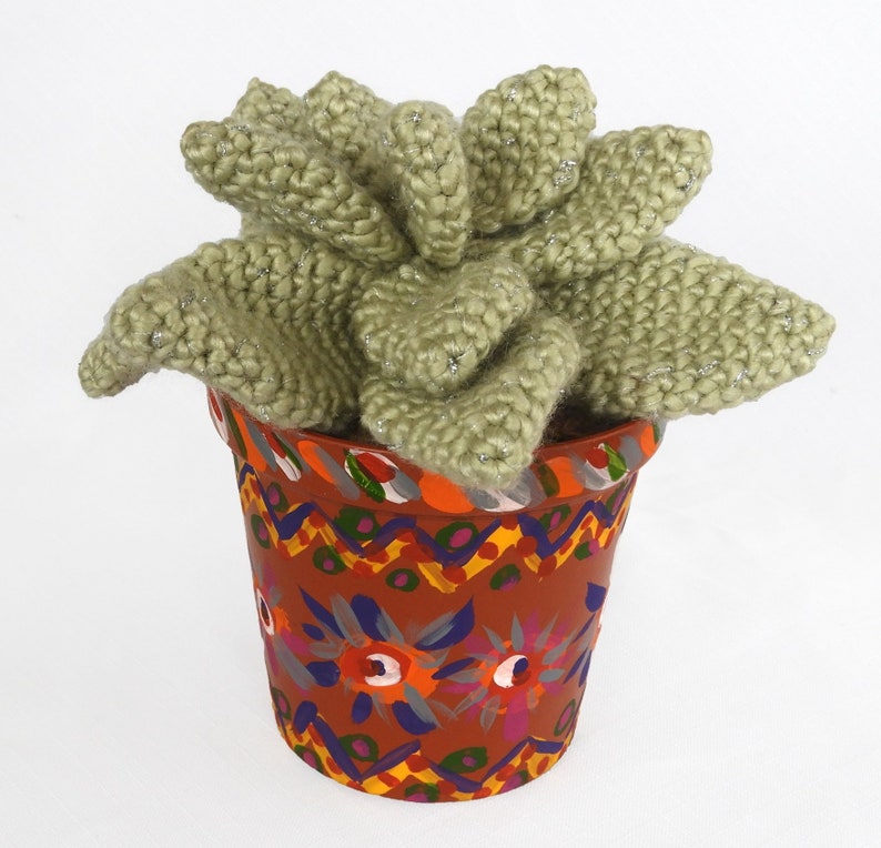 May include: A handmade, knitted succulent plant in a decorative terracotta pot. The plant is a soft green color with silver thread accents. The pot is painted with colorful floral and geometric patterns.