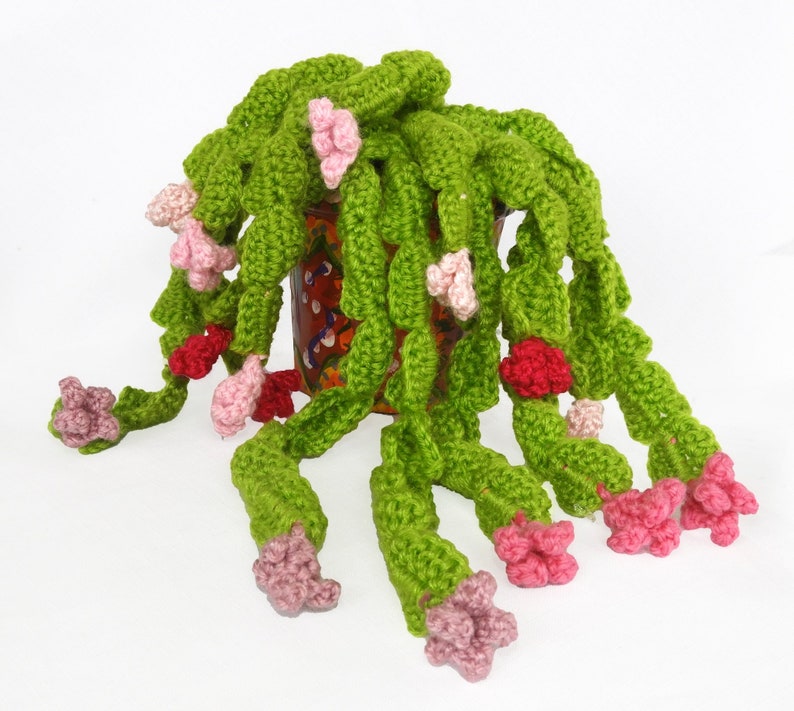 May include: A handmade crochet plant with trailing green leaves and pink and red flowers. The plant is in a decorative brown pot with colorful floral designs. The crochet work is detailed and the colors are vibrant.