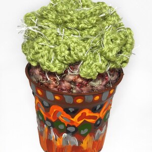 May include: A handmade, decorative cactus in a painted pot. The cactus is made of green yarn with white accents, and sits in a brown pot with colorful abstract designs in orange, yellow, gray, and white. The pot is approximately 4 inches tall.