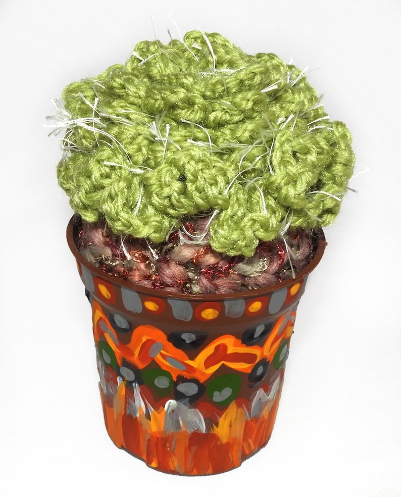 May include: A handmade, decorative cactus in a painted pot. The cactus is made of green yarn with white accents, and sits in a brown pot with colorful abstract designs in orange, yellow, gray, and white. The pot is approximately 4 inches tall.
