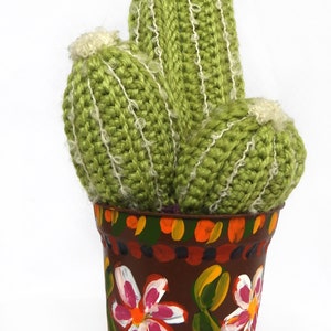 May include: A handmade, green crocheted cactus with white accents, potted in a brown, hand-painted pot adorned with colorful floral designs. The cactus has three stems and a fluffy top.