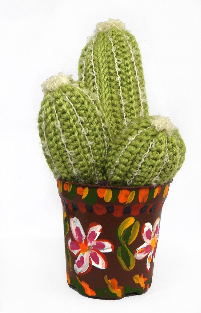 May include: A handmade, green crocheted cactus with white accents, potted in a brown, hand-painted pot adorned with colorful floral designs. The cactus has three stems and a fluffy top.