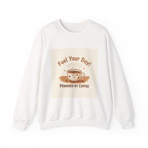 May include: White sweatshirt featuring a coffee cup graphic and the text "Fuel Your Day! Powered by Coffee." The coffee cup is brown with white polka dots and heart-shaped steam. The sweatshirt has long sleeves and a crew neck.
