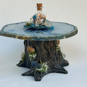 May include: A unique, handcrafted table with a blue-toned, round top. The table base is designed to resemble a tree trunk, adorned with seashells, faux flowers, and a glass bottle filled with shells and a cork stopper. The table is a decorative accent piece.