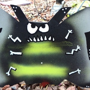 May include: A black and green bee-shaped sign with white accents. The sign reads "Beware of Bees" and has a cartoon bee face with large eyes and teeth.