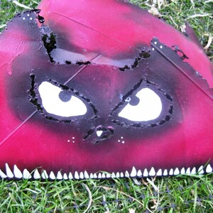 May include: A red and black painted metal object with a cat face and sharp teeth. The cat's eyes are white with black pupils.