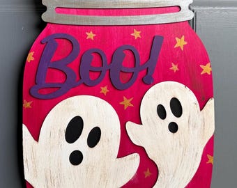 May include: A decorative wall hanging shaped like a mason jar with a pink background, featuring two white ghosts and the purple text "Boo!" across the top. The design includes small gold stars and a silver-painted lid with a jute twine hanger.
