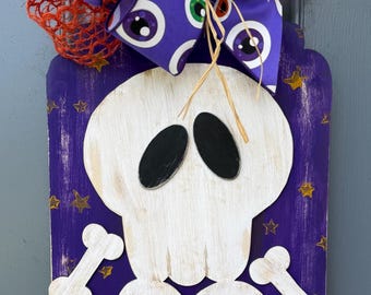 May include: A rectangular purple wooden wall hanging featuring a white skeleton with black eye sockets and rib cage details. The background has small gold stars. A large bow with an eyeball pattern and orange mesh netting sits at the top.